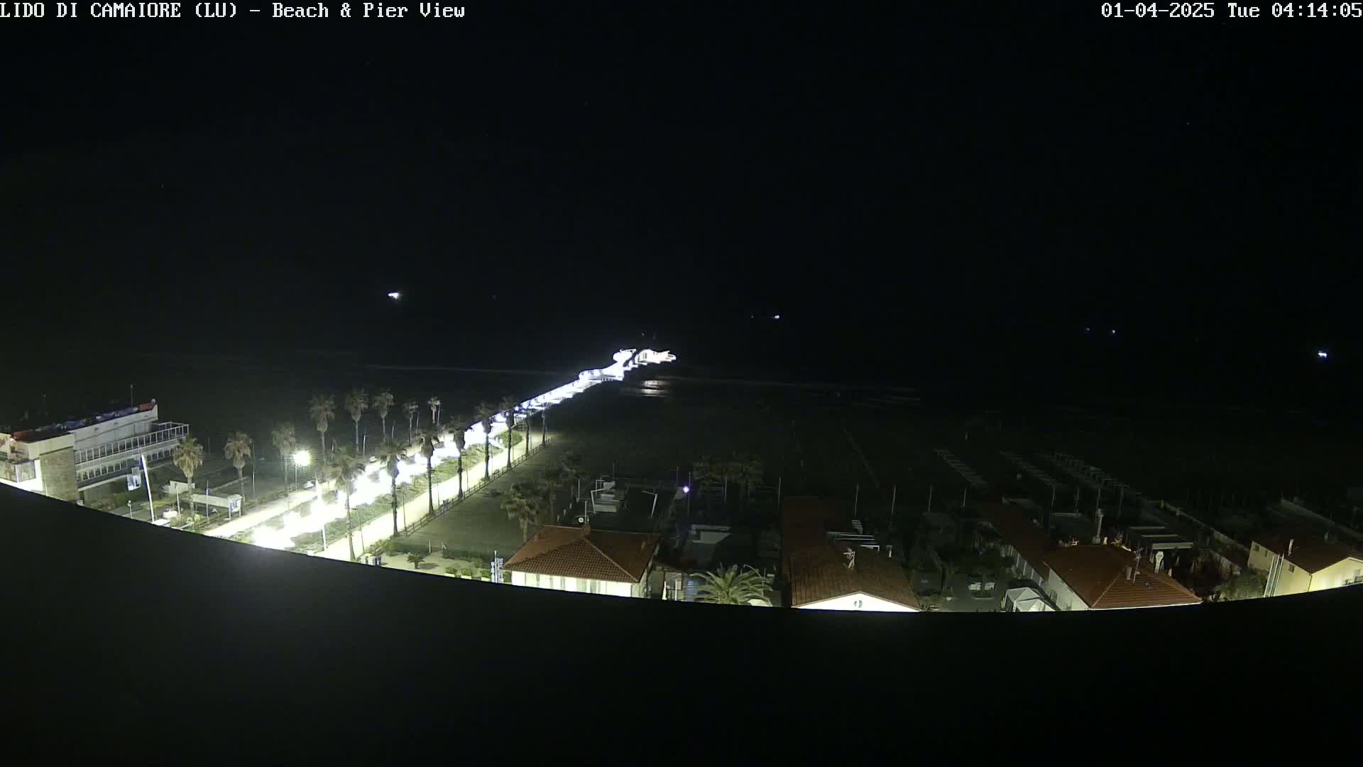 A nighttime, high-angle view shows a beach, pier, and seaside buildings illuminated by artificial light.