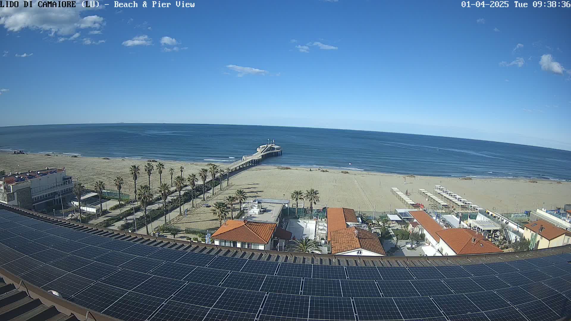 A sandy beach with a pier extends into a calm blue ocean under a mostly clear sky, viewed from above a building topped with solar panels.