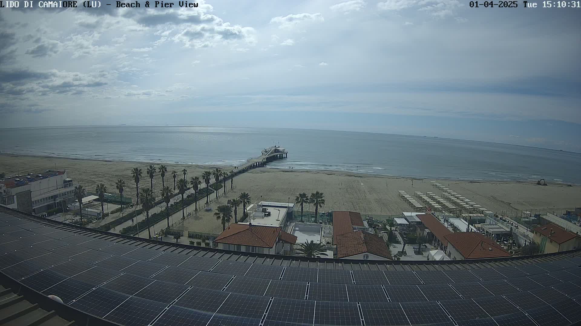 A mostly sunny day shows a sandy beach with a pier extending into a calm sea, viewed from above a rooftop covered in solar panels; palm trees line a walkway between the beach and buildings.