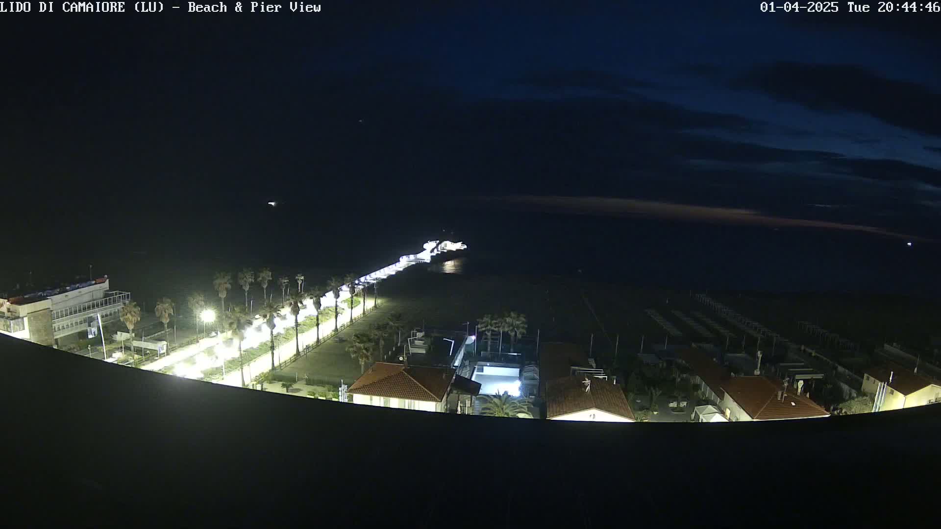 A nighttime, aerial view shows a beach, pier, and buildings illuminated by artificial lights under a dark sky.