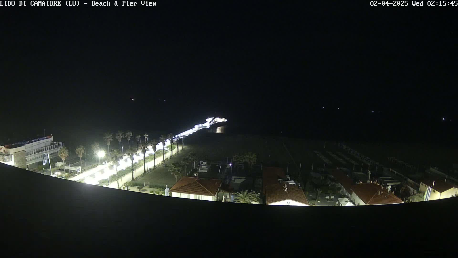 A nighttime, overhead view of a beach, pier, and buildings, illuminated by artificial light.