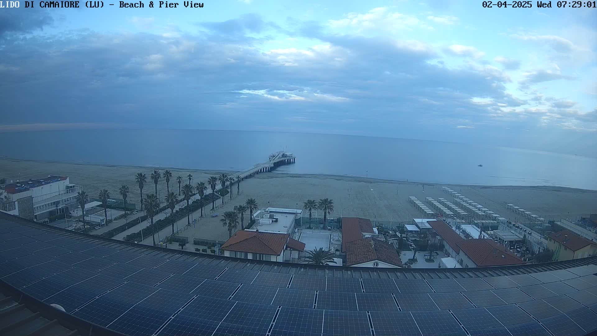 A mostly cloudy day reveals a beach scene with a pier extending into a calm sea, palm trees lining a beachfront walkway, buildings, and a large solar panel array on a roof in the foreground.
