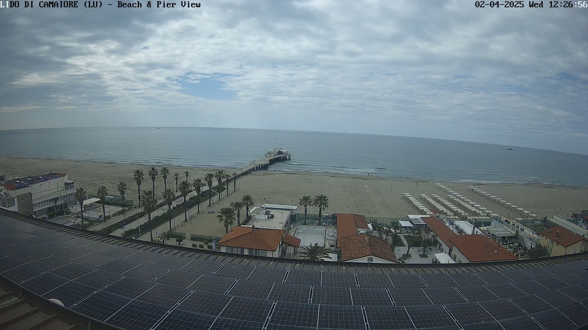 A mostly cloudy day reveals a sandy beach with palm trees, beach umbrellas, and a pier extending into a calm ocean, all viewed from above a rooftop covered in solar panels.