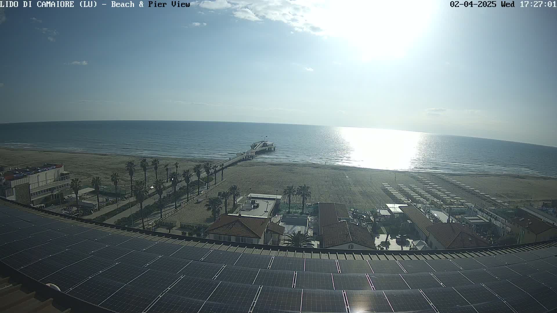 A sunny day shows a beach with a pier, palm trees, and buildings, viewed from a rooftop covered in solar panels.