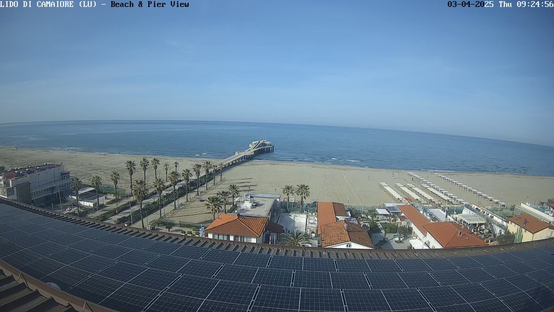 A sunny day overlooking a sandy beach, a pier extending into a calm ocean, palm trees lining the beachfront, and buildings with solar panels on the roof.