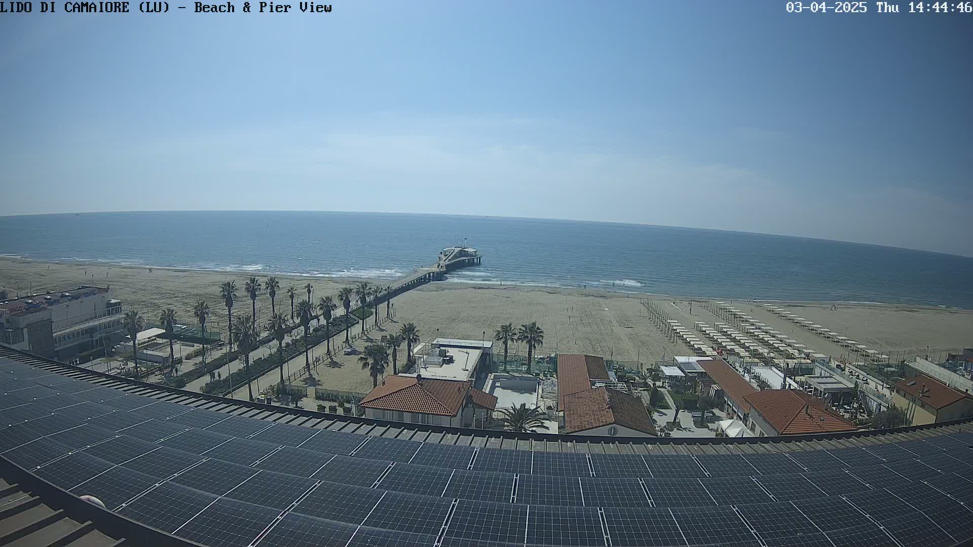 A mostly sunny day overlooking a beach with a pier, palm trees, beach umbrellas, buildings, and a large solar panel array on a rooftop.