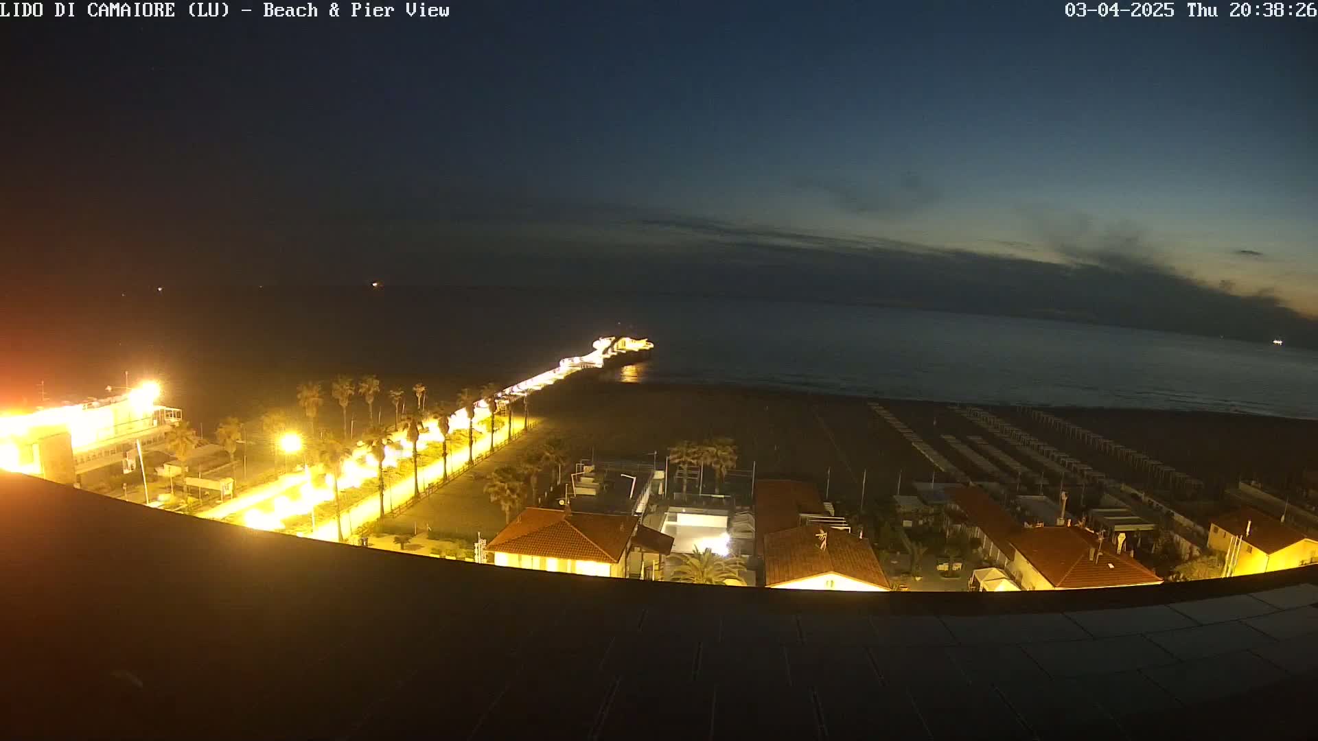A nighttime view of a beach and pier, illuminated by streetlights, with a calm, dark sea under a mostly clear sky.
