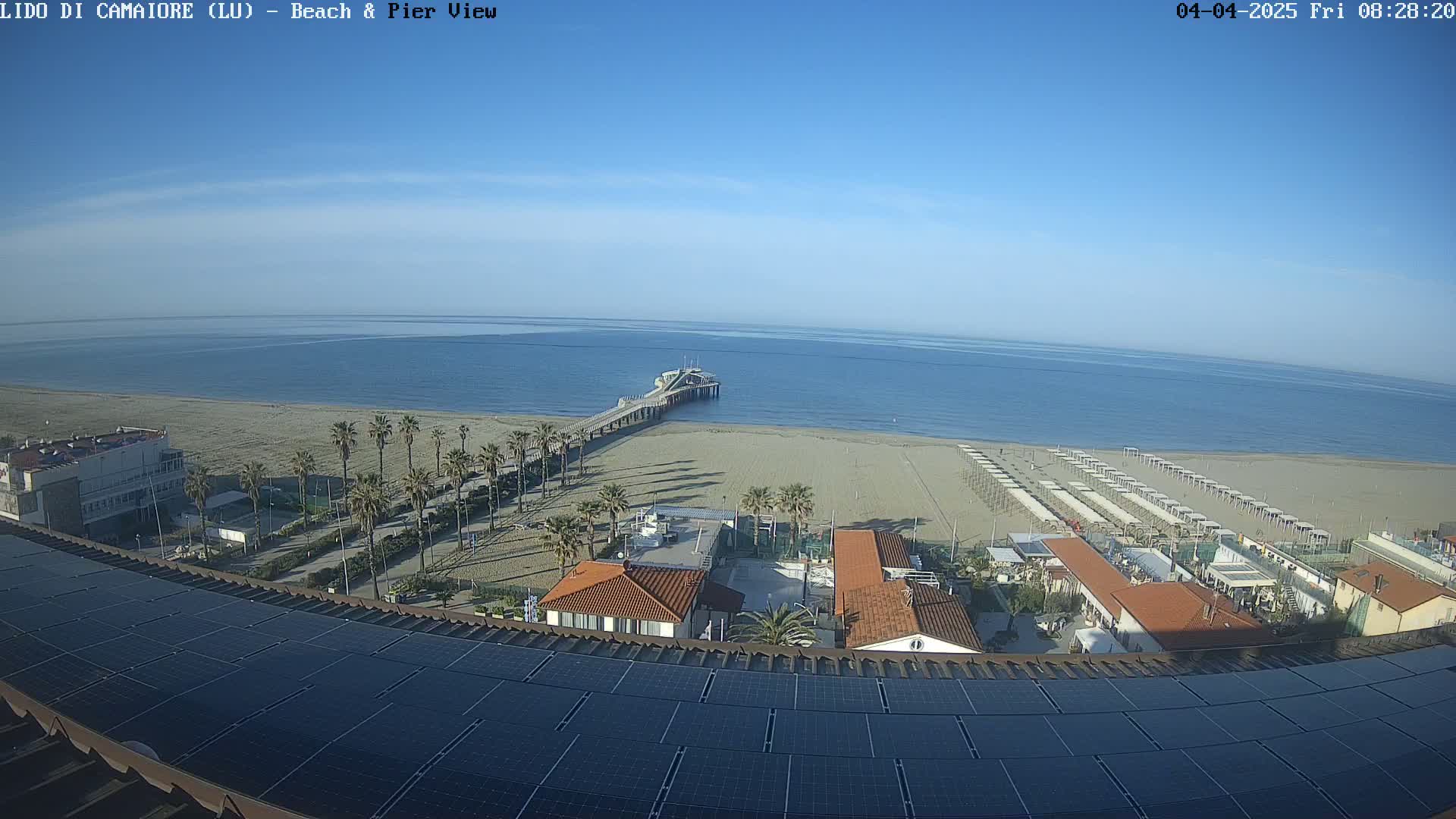 A beach with a pier, palm trees, and beach umbrellas is visible under a clear blue sky, viewed from above a building with solar panels on its roof.