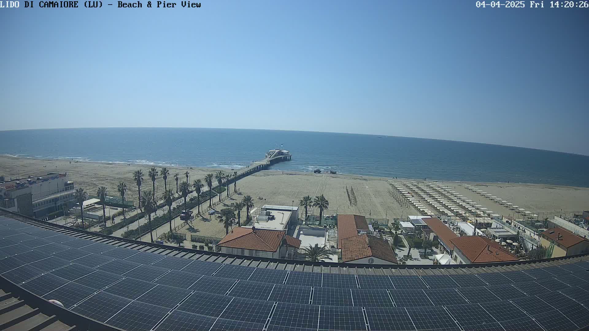A sunny beach scene is viewed from above, showing a pier extending into a calm ocean, rows of beach umbrellas, palm trees, and buildings, all situated atop a large solar panel array.