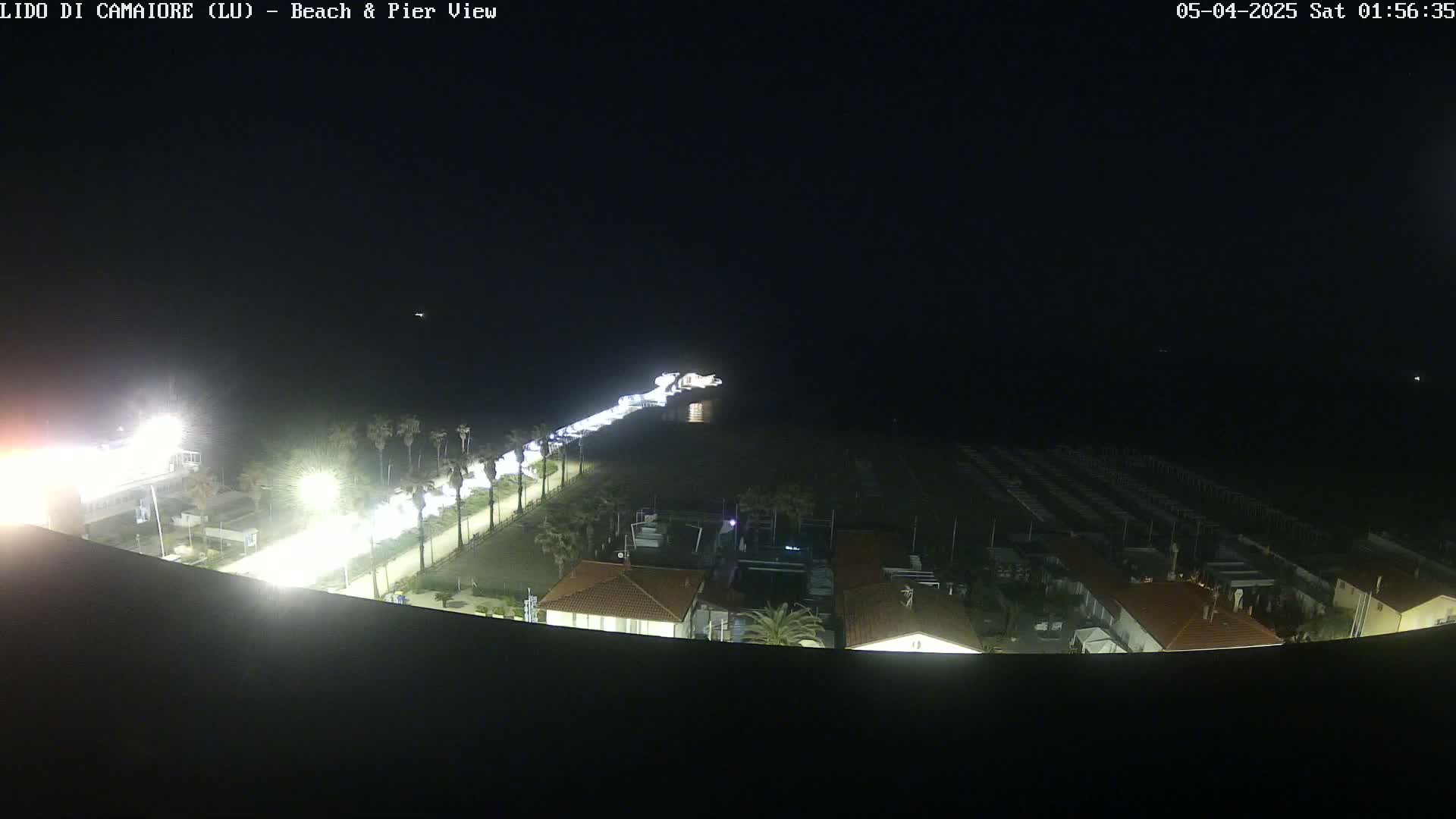 A nighttime, high-angle view shows a beach, pier, and beachfront buildings illuminated by artificial light.
