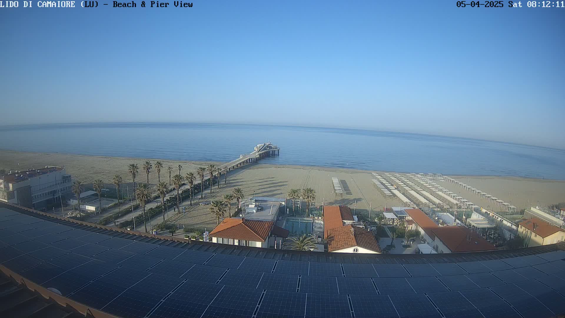 A clear, sunny day overlooking a beach with rows of beach umbrellas, palm trees, a pier extending into a calm ocean, and a building topped with solar panels.