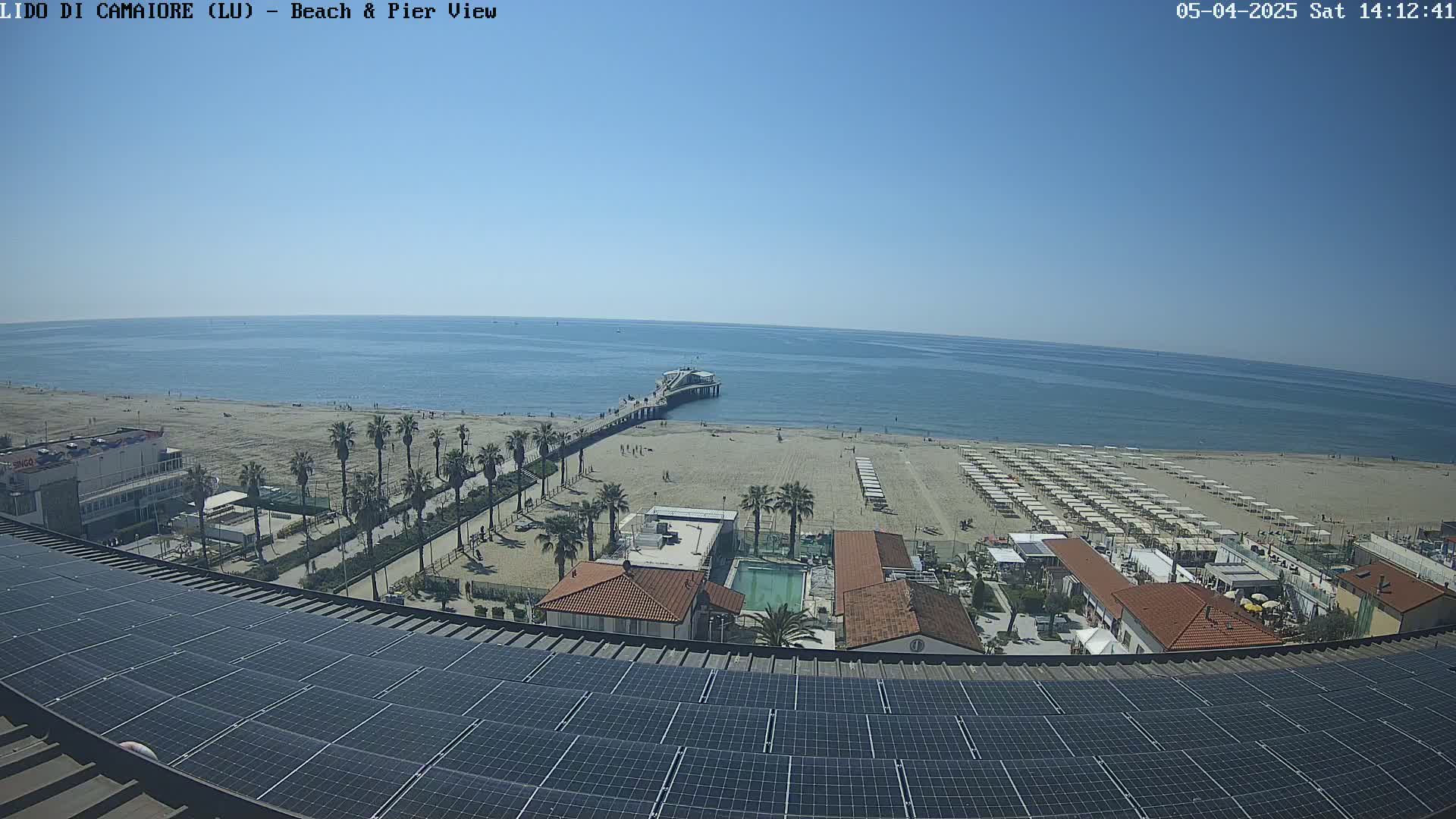 A beach scene on a sunny day is viewed from above a rooftop covered in solar panels; a pier extends into a calm ocean, and beach umbrellas are set up on the sand.