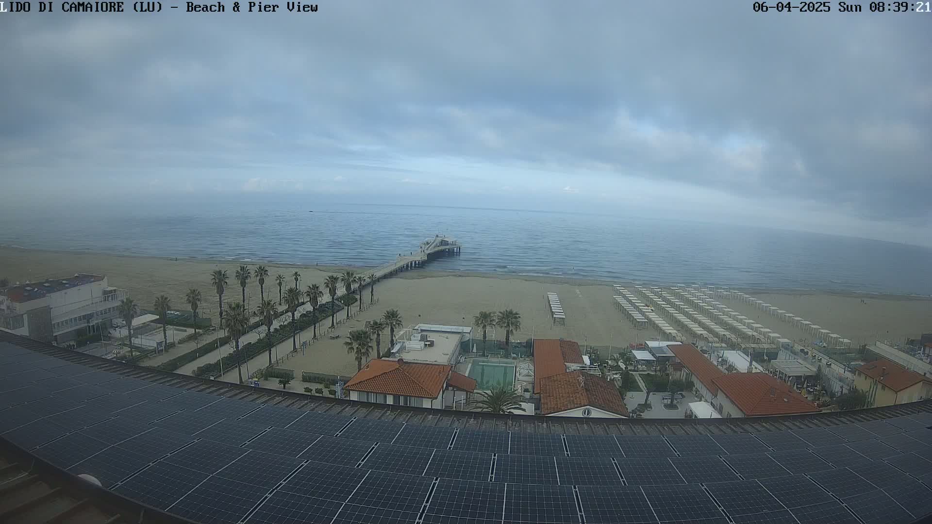 A beach with a pier, lined with beach umbrellas, is visible from a rooftop with solar panels under an overcast sky.