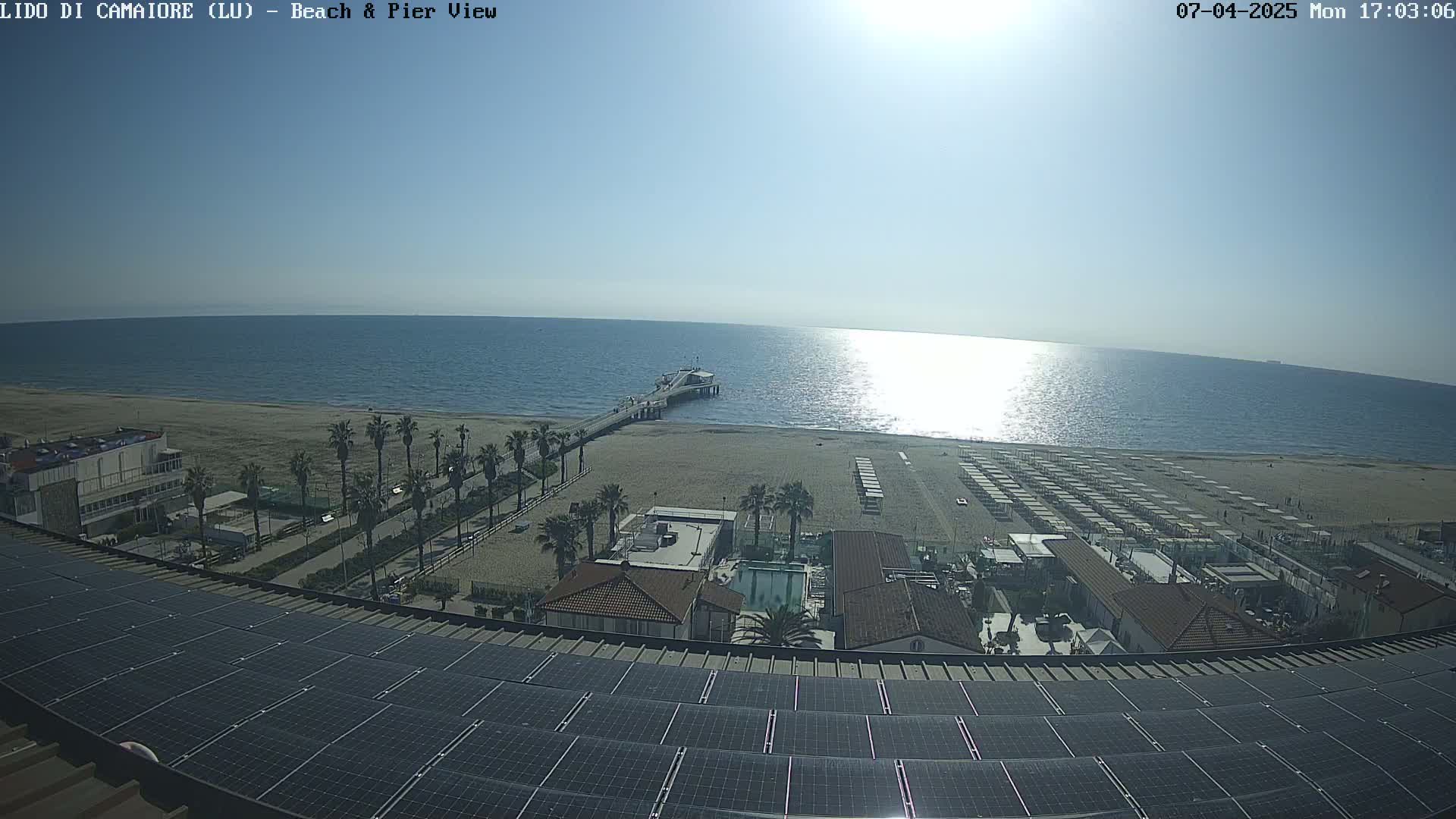 A sunny day at a beach shows a pier extending into the calm ocean, with rows of beach umbrellas and buildings visible from a rooftop with solar panels.