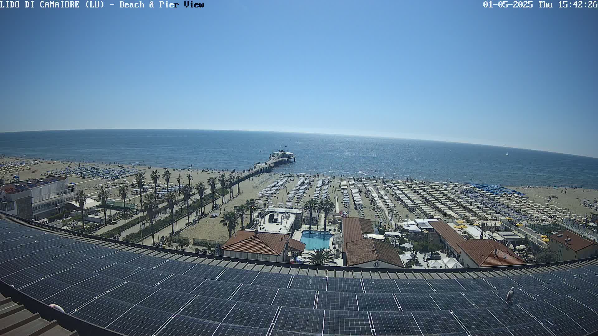 A sunny beach scene with numerous beach umbrellas, a pier extending into a calm ocean, and a rooftop covered in solar panels.