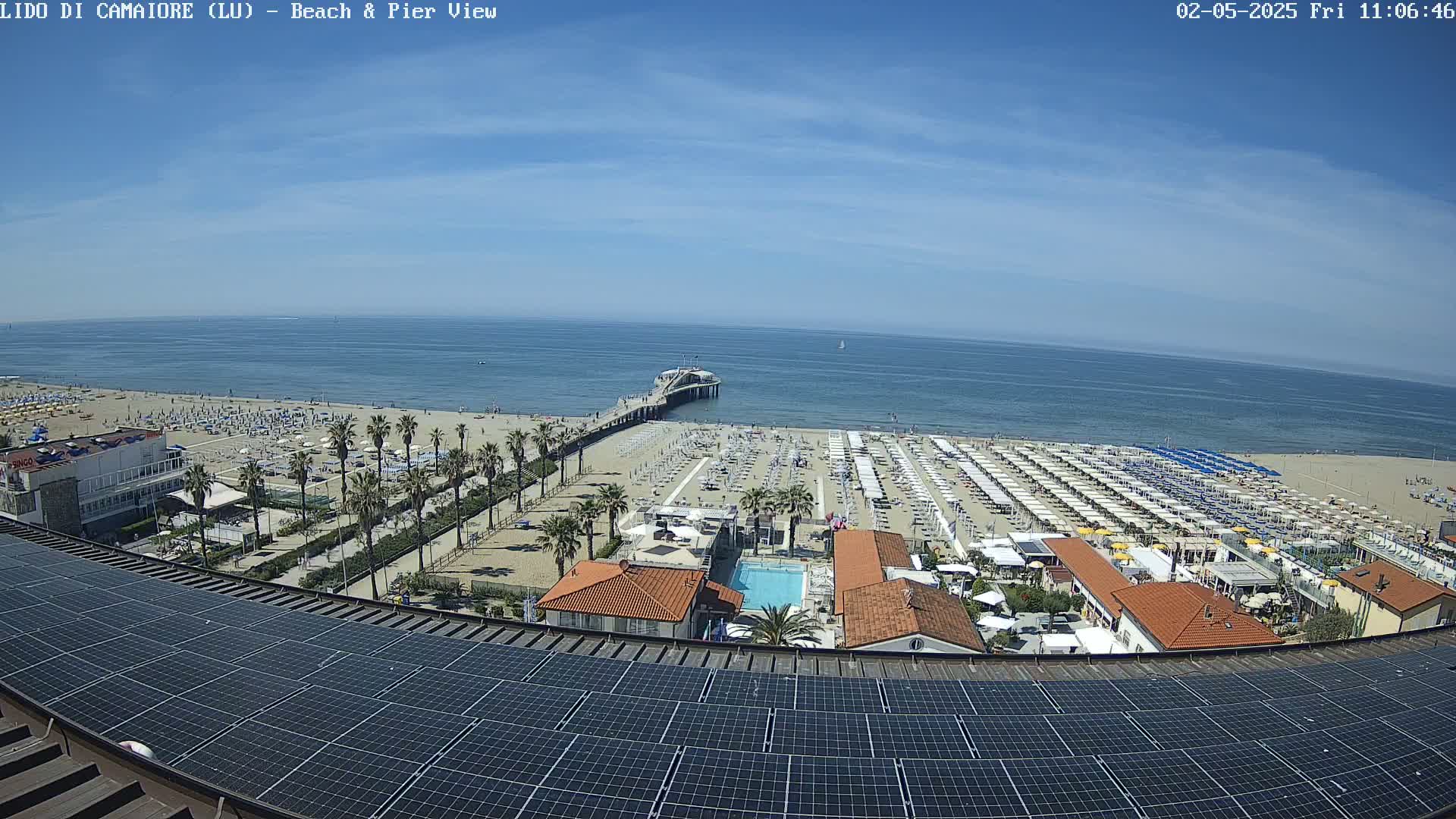 A sandy beach with numerous beach umbrellas, a pier extending into a calm blue sea under a sunny sky, is viewed from above a building with solar panels on its roof.