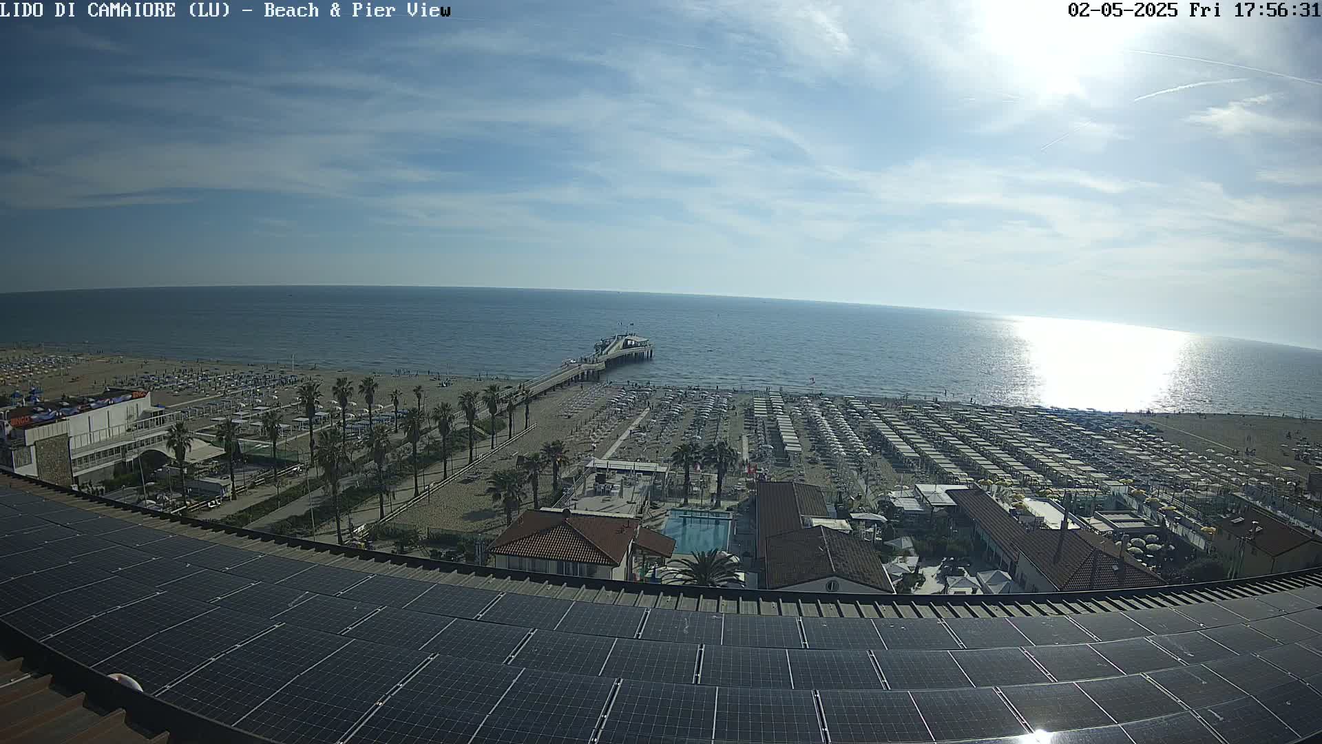 A sunny day overlooking a beach with many umbrellas, palm trees, a pier, and a building with solar panels on its roof.