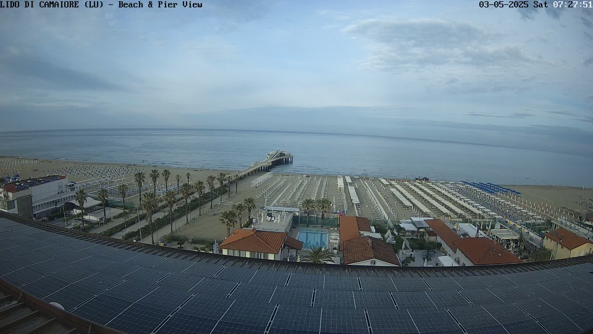 An aerial view shows a beach with rows of umbrellas and lounge chairs, a pier extending into a calm sea under a partly cloudy sky.