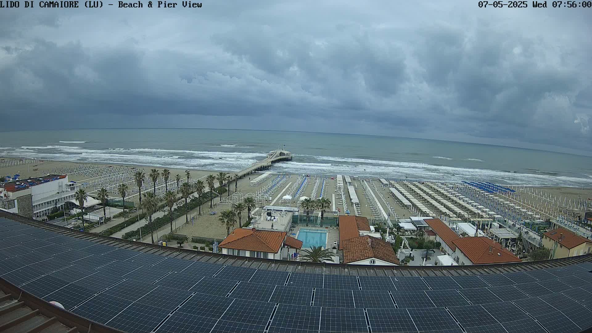 A beach with rows of beach umbrellas and a pier extends into a wavy ocean under a cloudy sky.