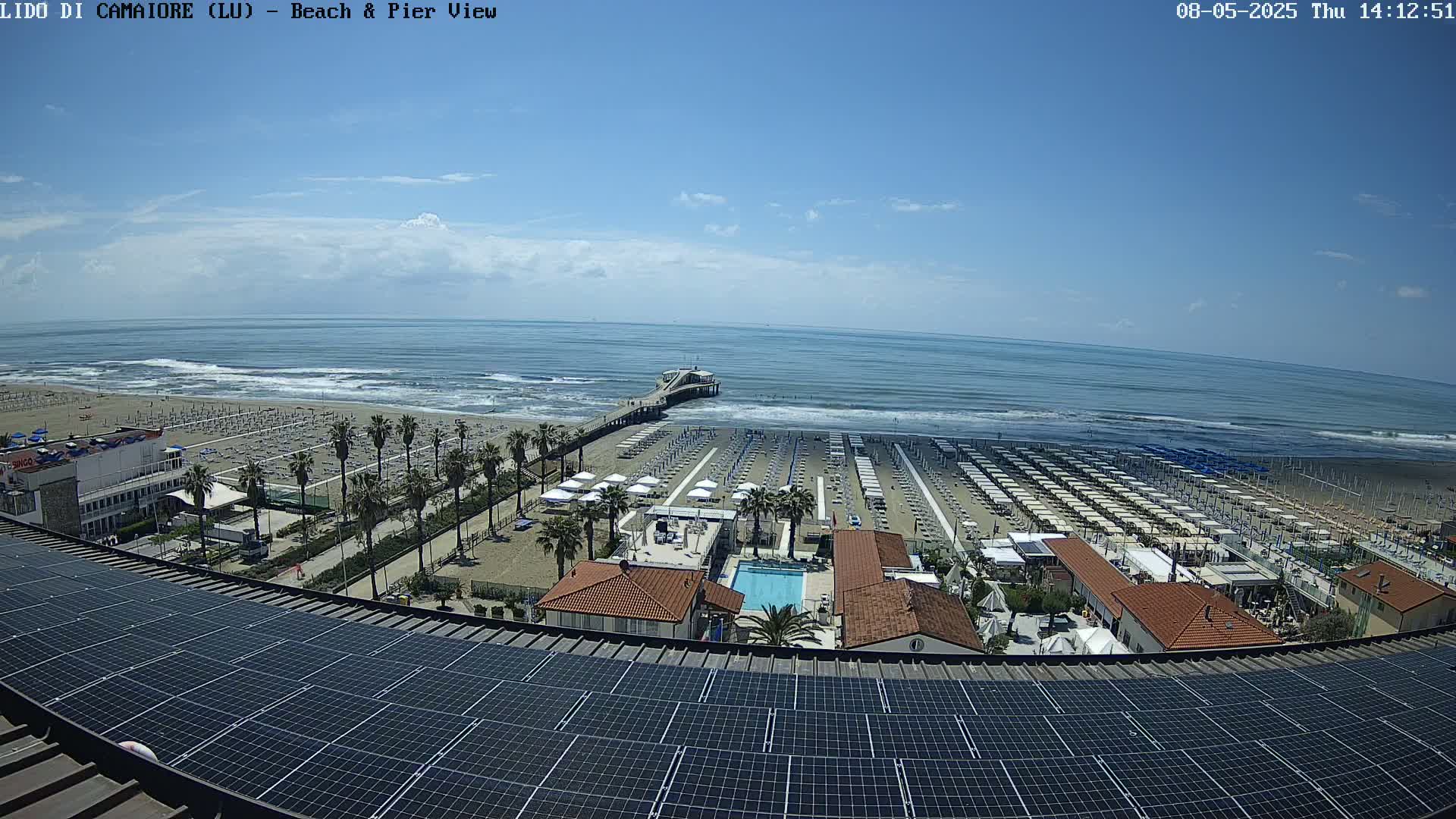 A beach with rows of umbrellas and a pier extending into a mostly calm ocean under a partly cloudy sky is viewed from above a rooftop covered in solar panels.