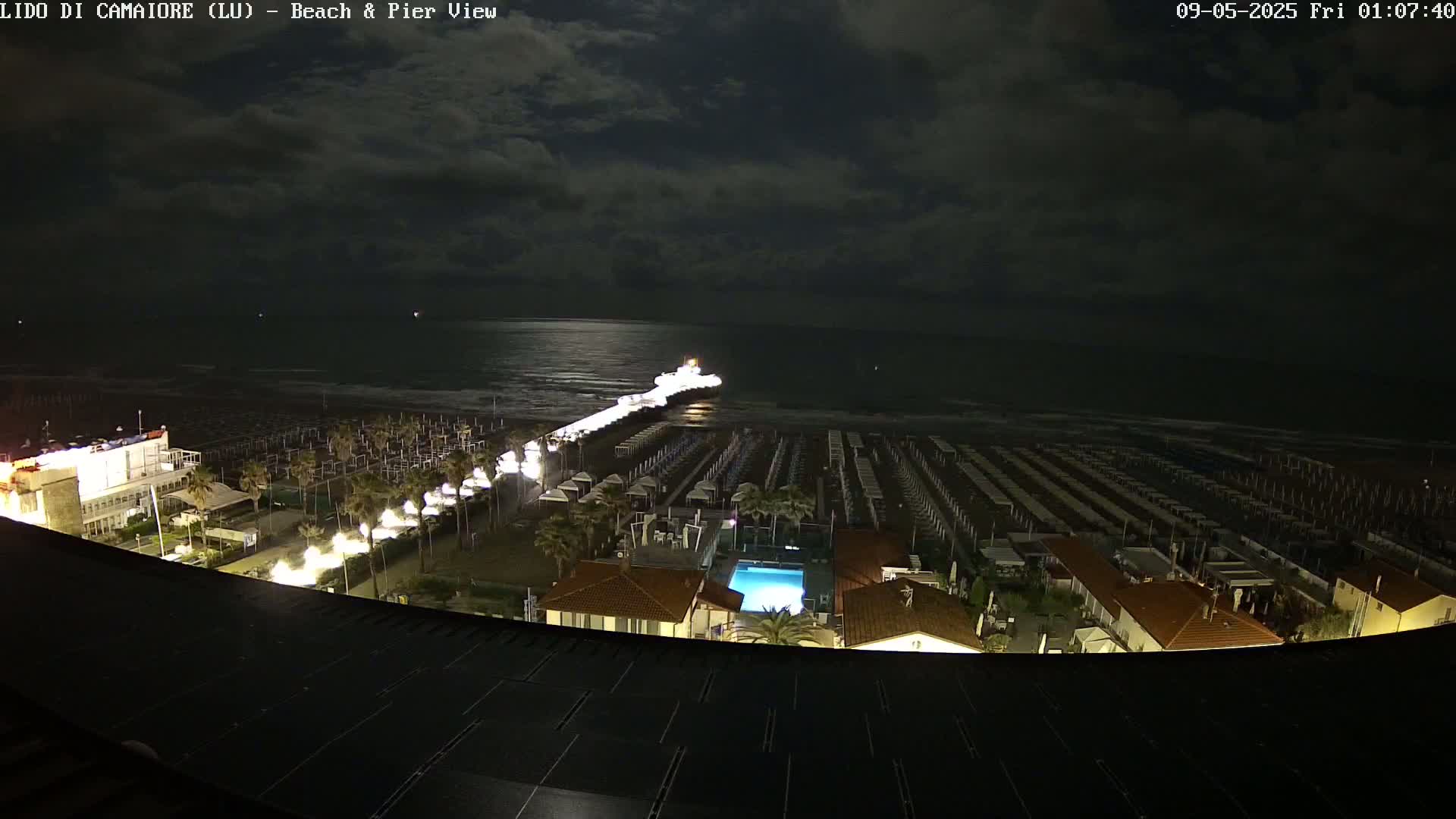 A nighttime view of a beach with rows of beach umbrellas, a pier extending into a calm sea under a partly cloudy sky.