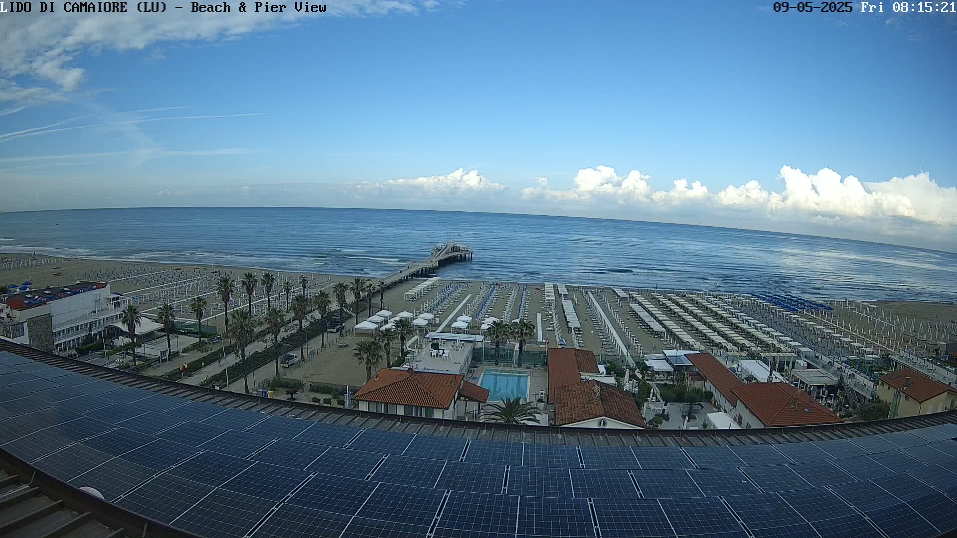 A partly cloudy day shows a beach with rows of umbrellas and a pier extending into a calm ocean, viewed from a rooftop with solar panels.
