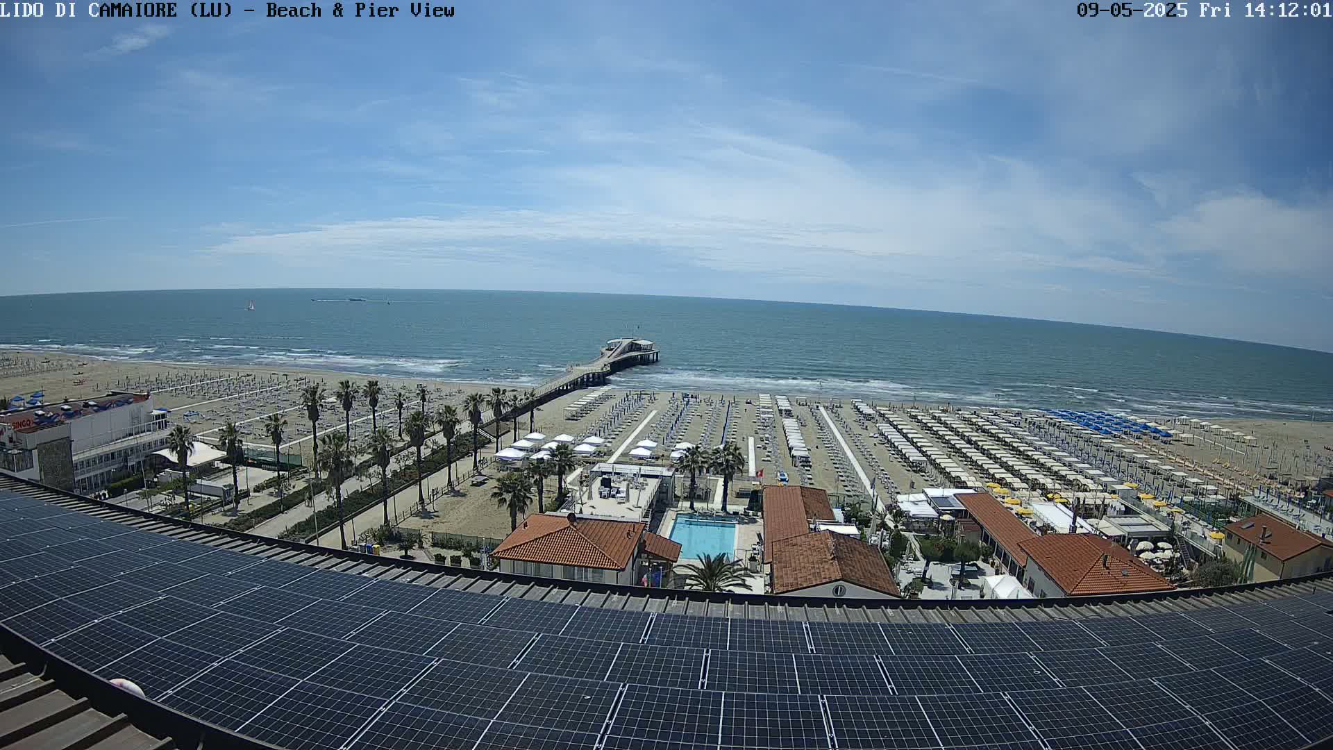 A sunny day at a beach resort shows a pier extending into a calm ocean, with numerous beach umbrellas and lounge chairs arranged on the sand, viewed from above a rooftop with solar panels.