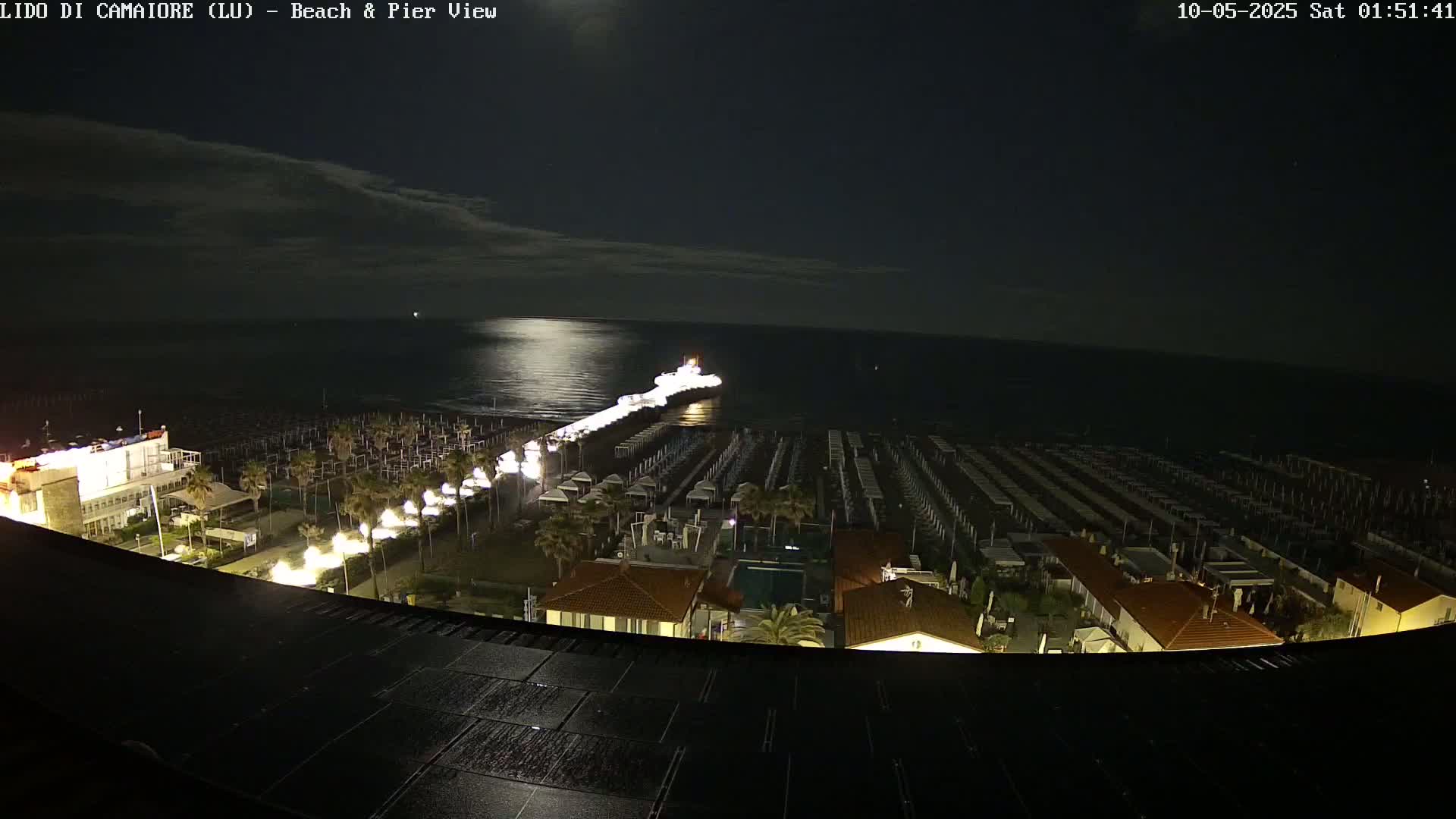 A nighttime, high-angle view shows a beach with rows of closed beach umbrellas, a pier extending into a calm sea under a moonlit sky.