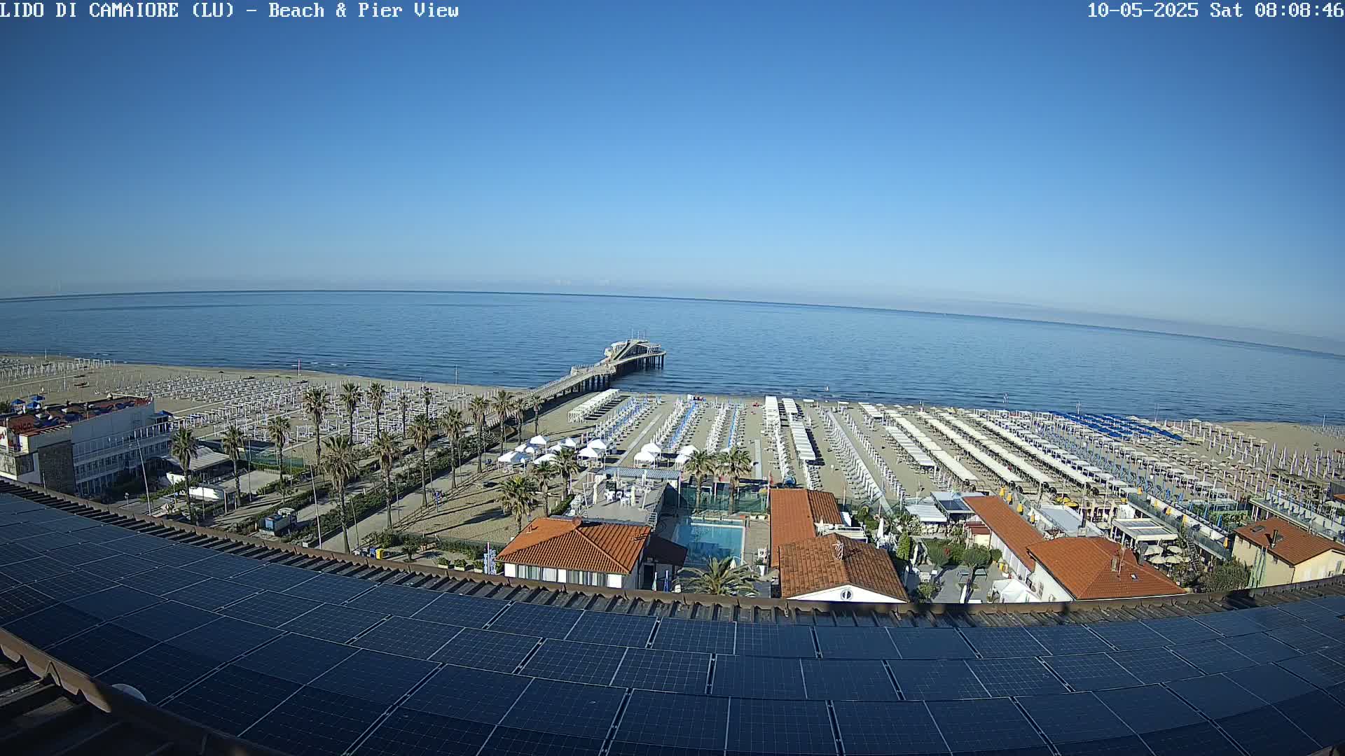 A beach with rows of umbrellas and a pier extends into a calm, blue ocean under a clear, sunny sky, viewed from a rooftop with solar panels.