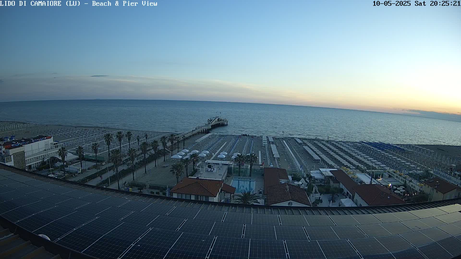 A beach with rows of beach umbrellas and a pier extends into a calm, twilight ocean viewed from a rooftop with solar panels.