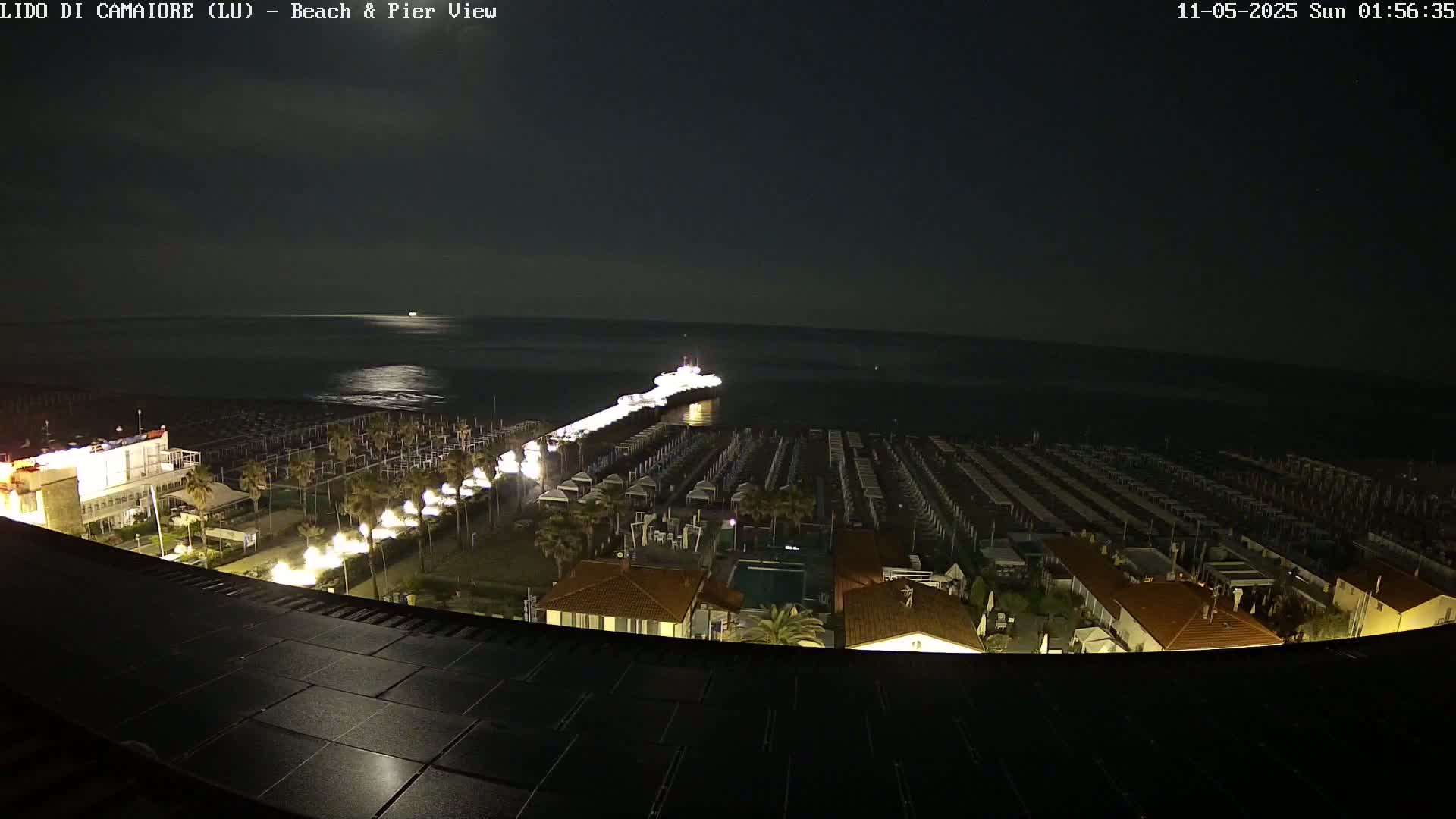 A nighttime aerial view of a beach and pier, with rows of beach umbrellas illuminated, a calm sea, and a vessel at the end of the pier.
