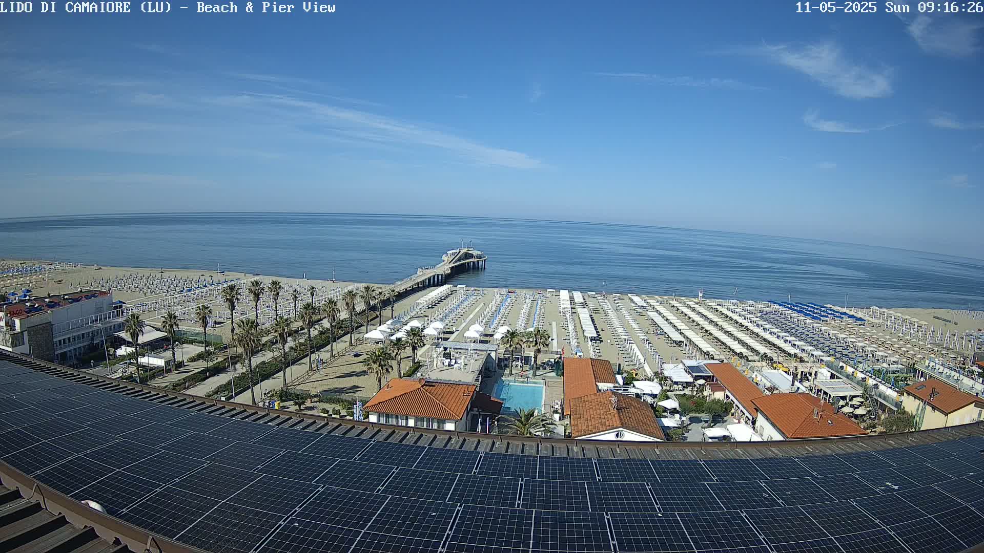 A beach with rows of umbrellas and a pier extends into a calm blue sea under a clear sky.