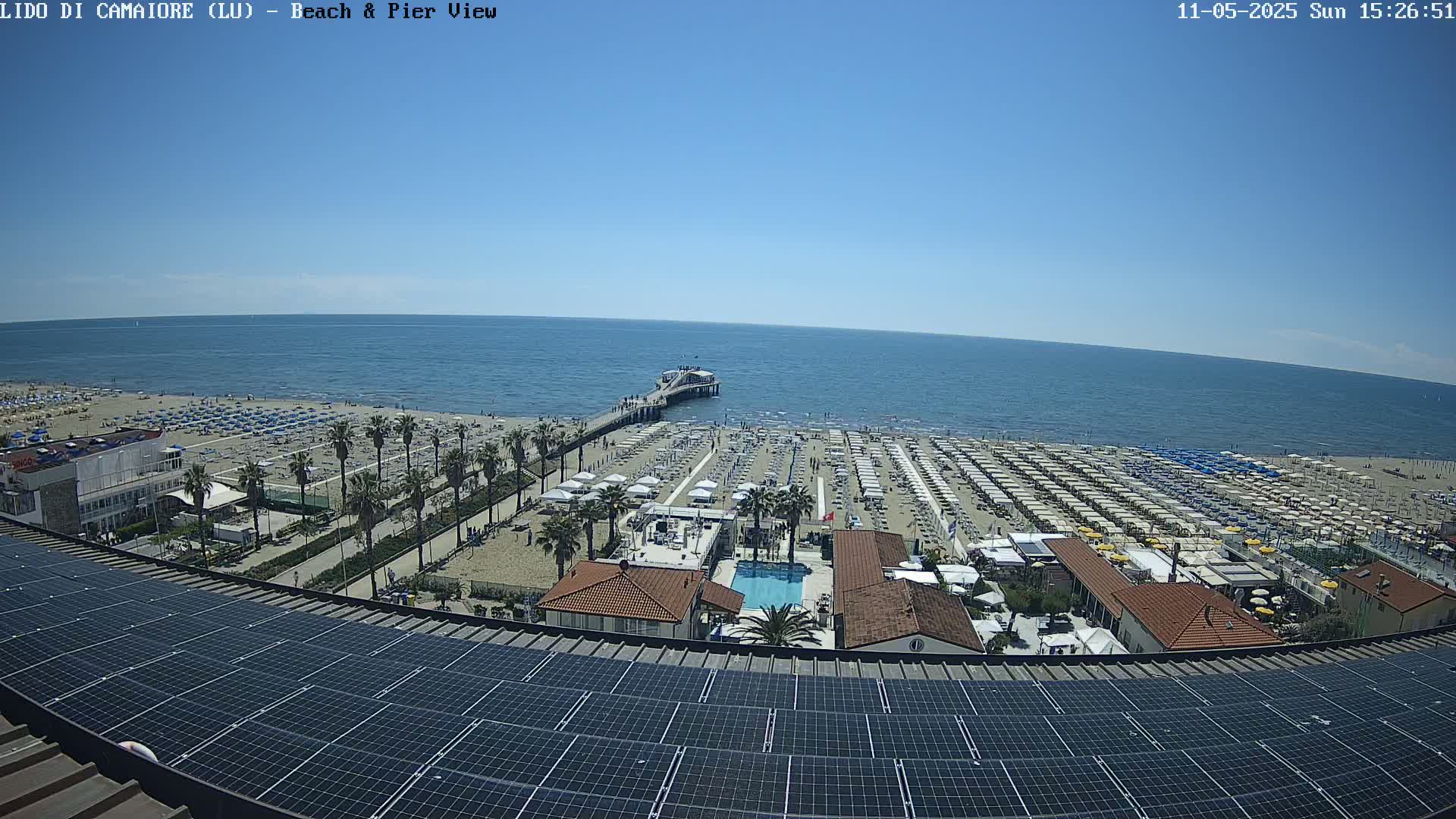 A sunny day at a beach with a pier, numerous beach umbrellas, and a building with a solar panel array on its roof.