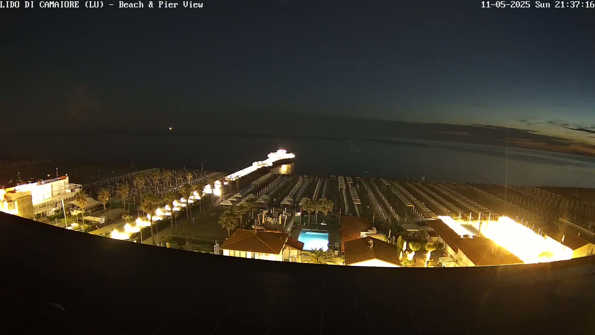 A nighttime, aerial view shows a pier extending into a dark body of water, with brightly lit beach umbrellas and buildings lining the shore.