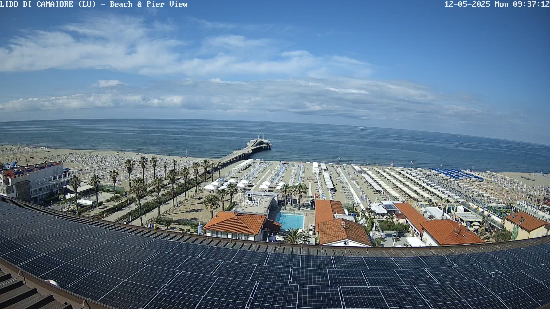 A beach with numerous umbrellas and a pier extends into a calm, partly cloudy ocean, viewed from above a rooftop covered in solar panels.