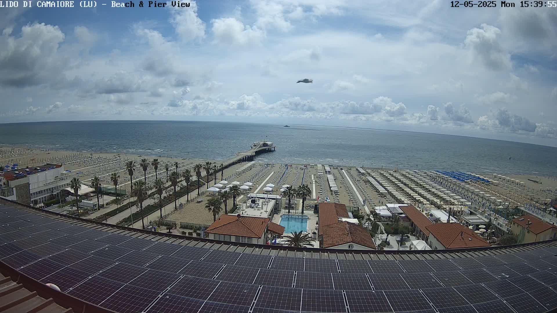 A partly cloudy day shows a beach with rows of umbrellas, a pier, a swimming pool, and palm trees, all viewed from above a rooftop covered in solar panels.