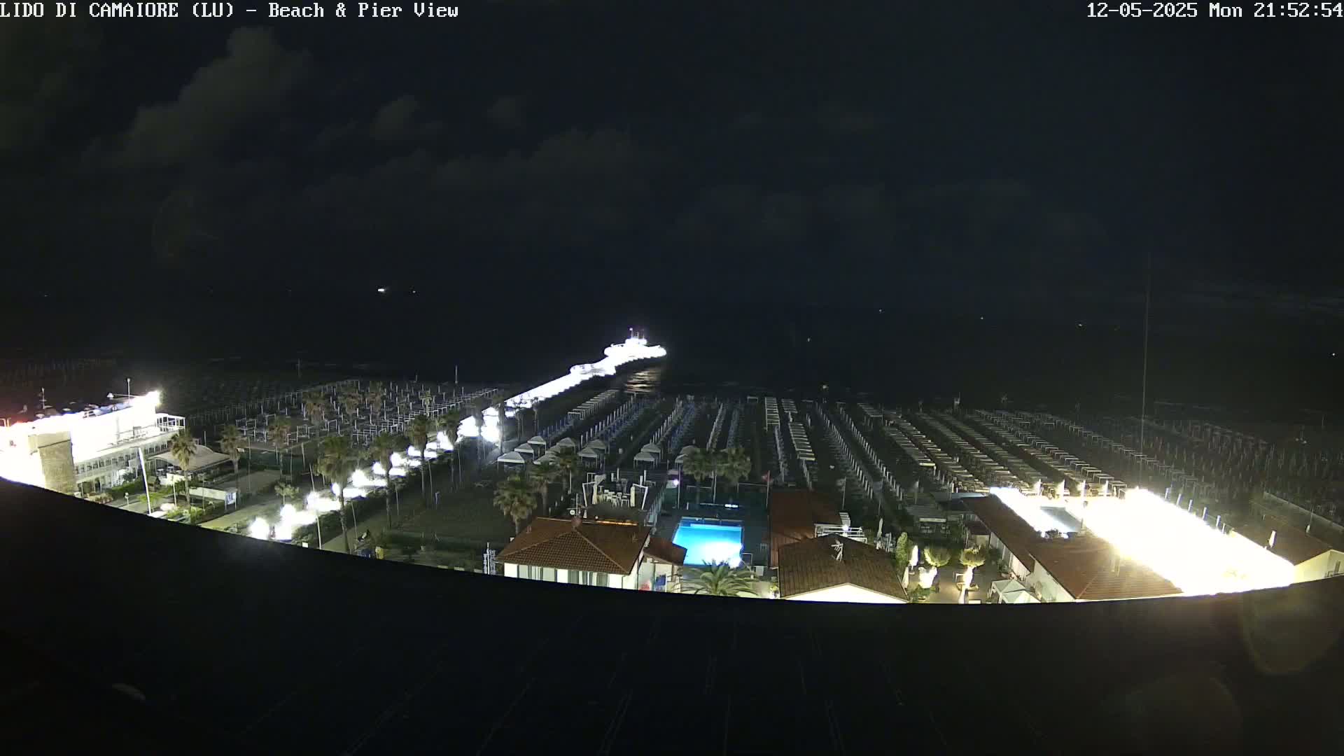 A nighttime aerial view shows a beach with rows of closed beach umbrellas, a pier with a boat at the end, and a building with a lit-up swimming pool under a partly cloudy sky.