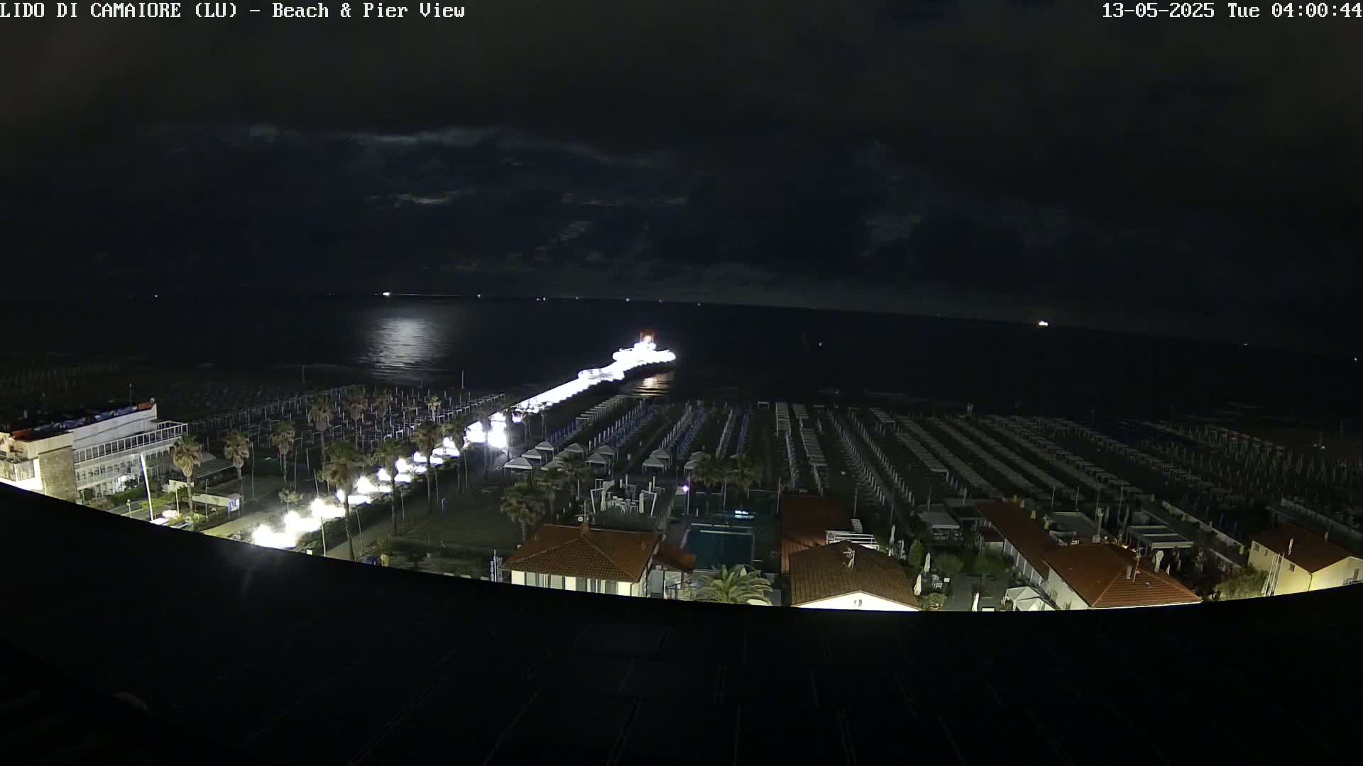 A nighttime, high-angle view of a beach with rows of closed beach umbrellas, a pier extending into a calm sea, and buildings illuminated under a partly cloudy sky.