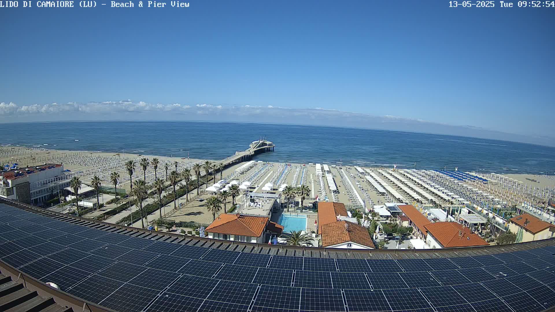 A beach with rows of umbrellas and lounge chairs, a pier extending into a calm blue ocean under a mostly clear sky, is viewed from above a rooftop covered in solar panels.