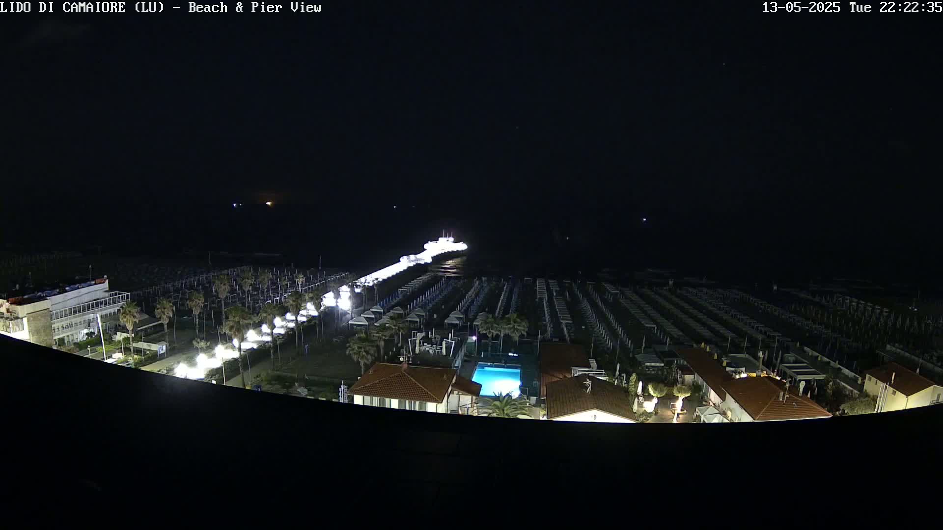 A nighttime aerial view shows a beach with rows of beach umbrellas and a pier extending into dark water, with a building and swimming pool visible nearby.