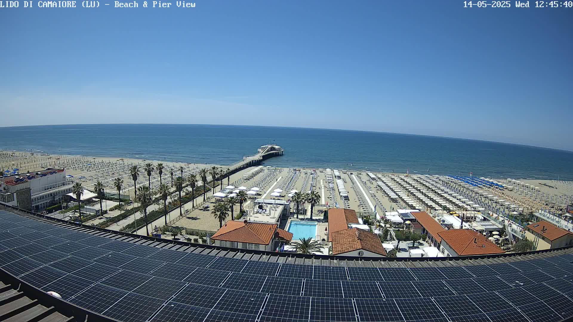 A sunny day at a beach with numerous umbrellas and lounge chairs, a pier extending into the calm ocean, and a solar panel array on a rooftop overlooking the scene.