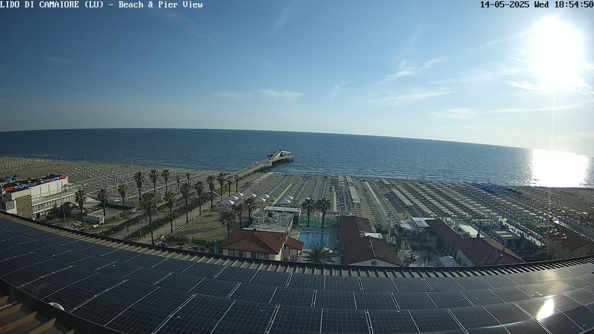 A sunny day at a beach with numerous beach umbrellas, a pier extending into a calm ocean, and solar panels on a rooftop in the foreground.