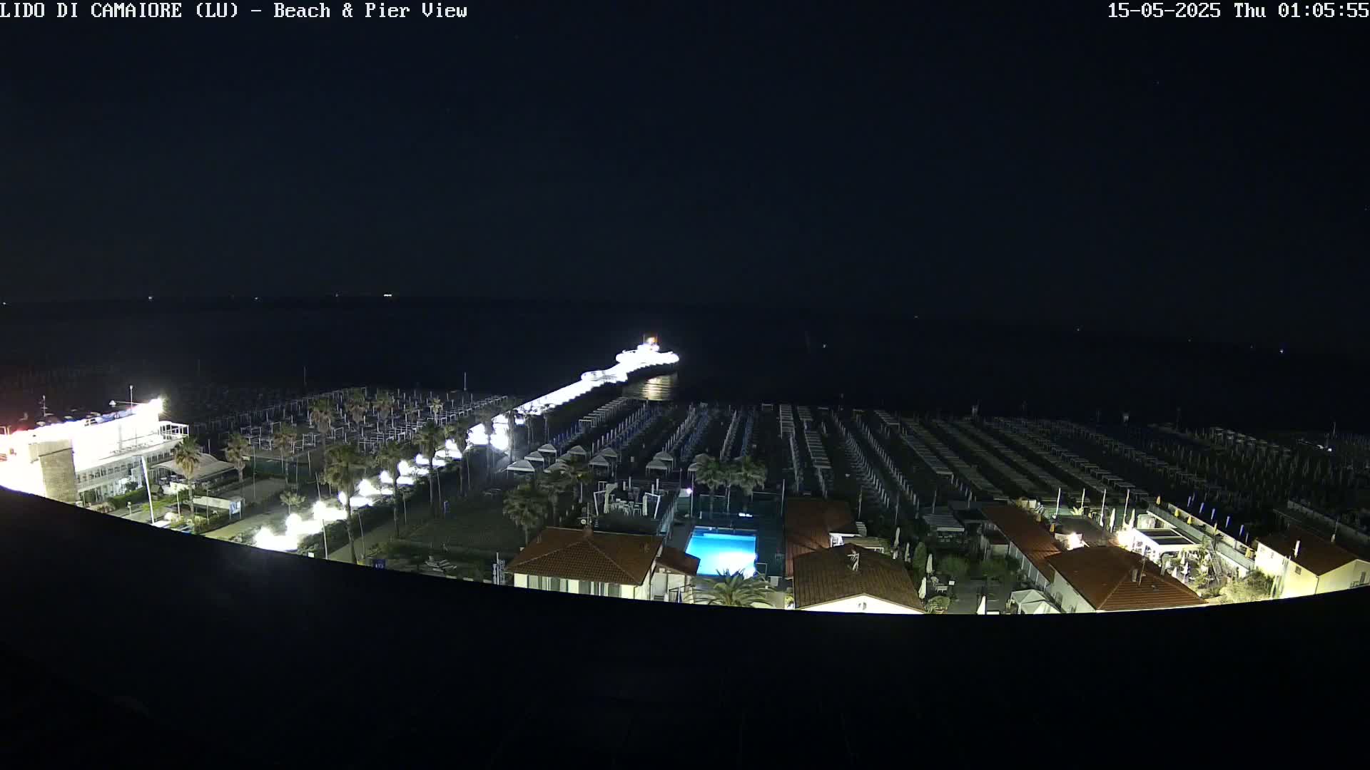A nighttime, aerial view shows a pier extending into a dark body of water, with a beach lined with rows of beach umbrellas and a building with a swimming pool.