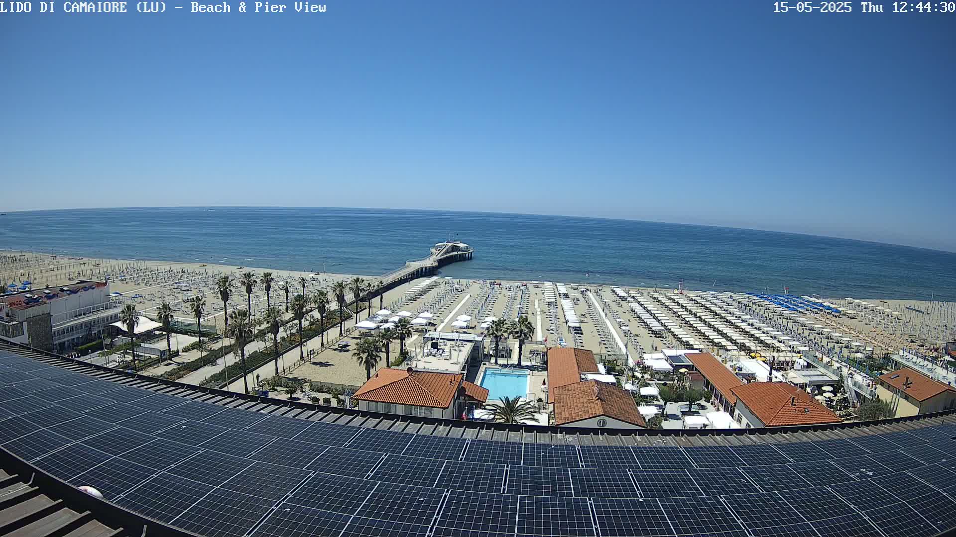 A beach with numerous umbrellas and a pier extends into a calm, blue ocean under a clear sky, viewed from above a rooftop covered in solar panels.