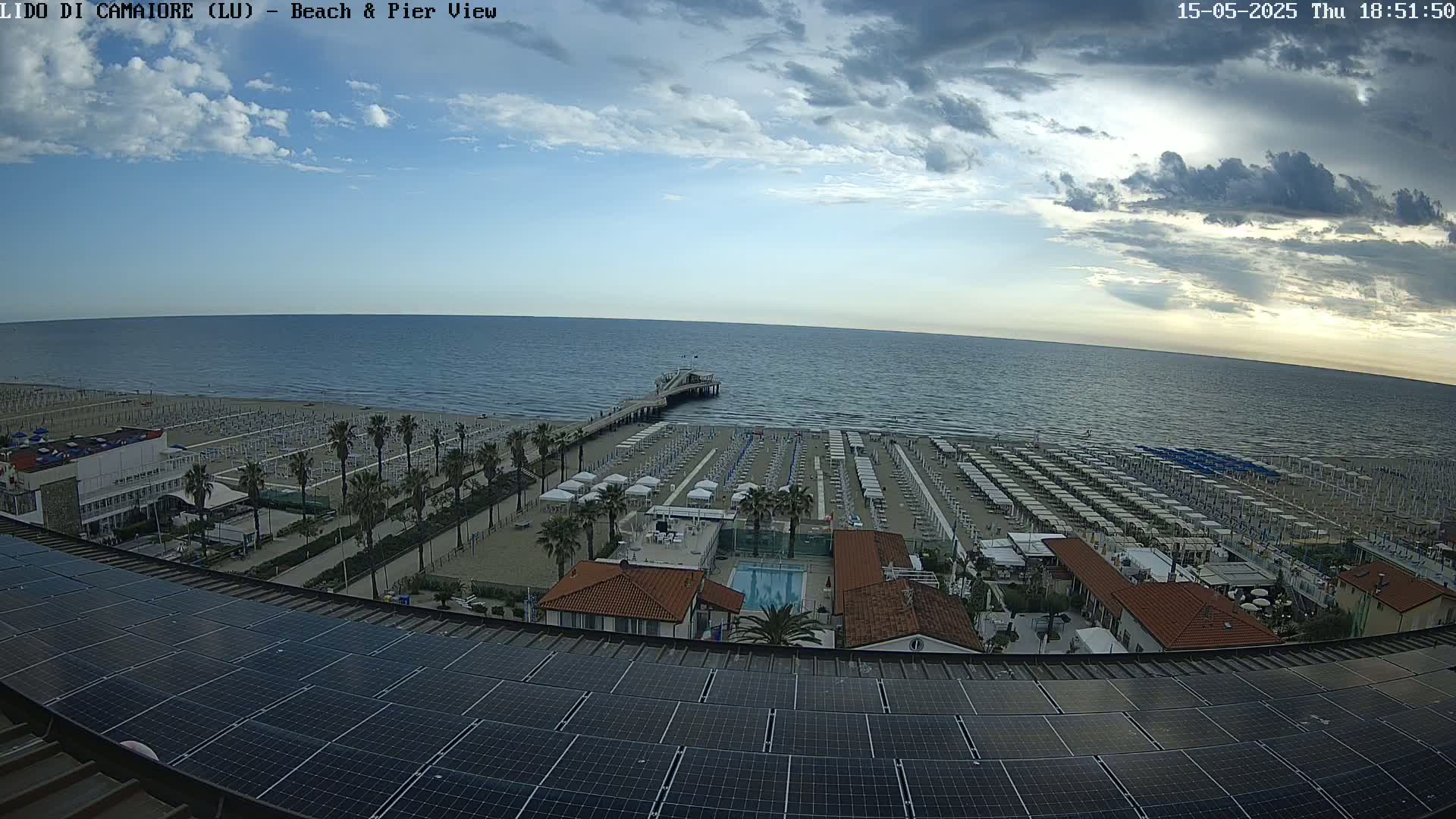 A beach with rows of umbrellas and a pier extends into a calm sea under a partly cloudy sky.