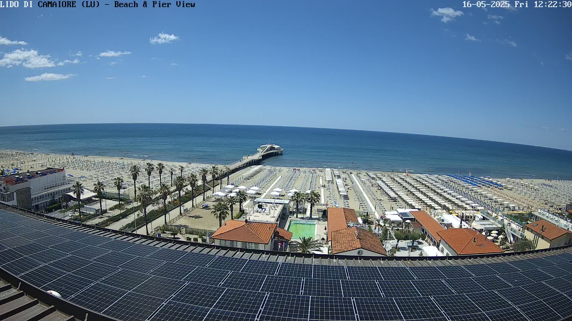 A sunny day overlooks a beach with numerous umbrellas and a pier extending into a calm ocean, viewed from above a rooftop covered in solar panels.
