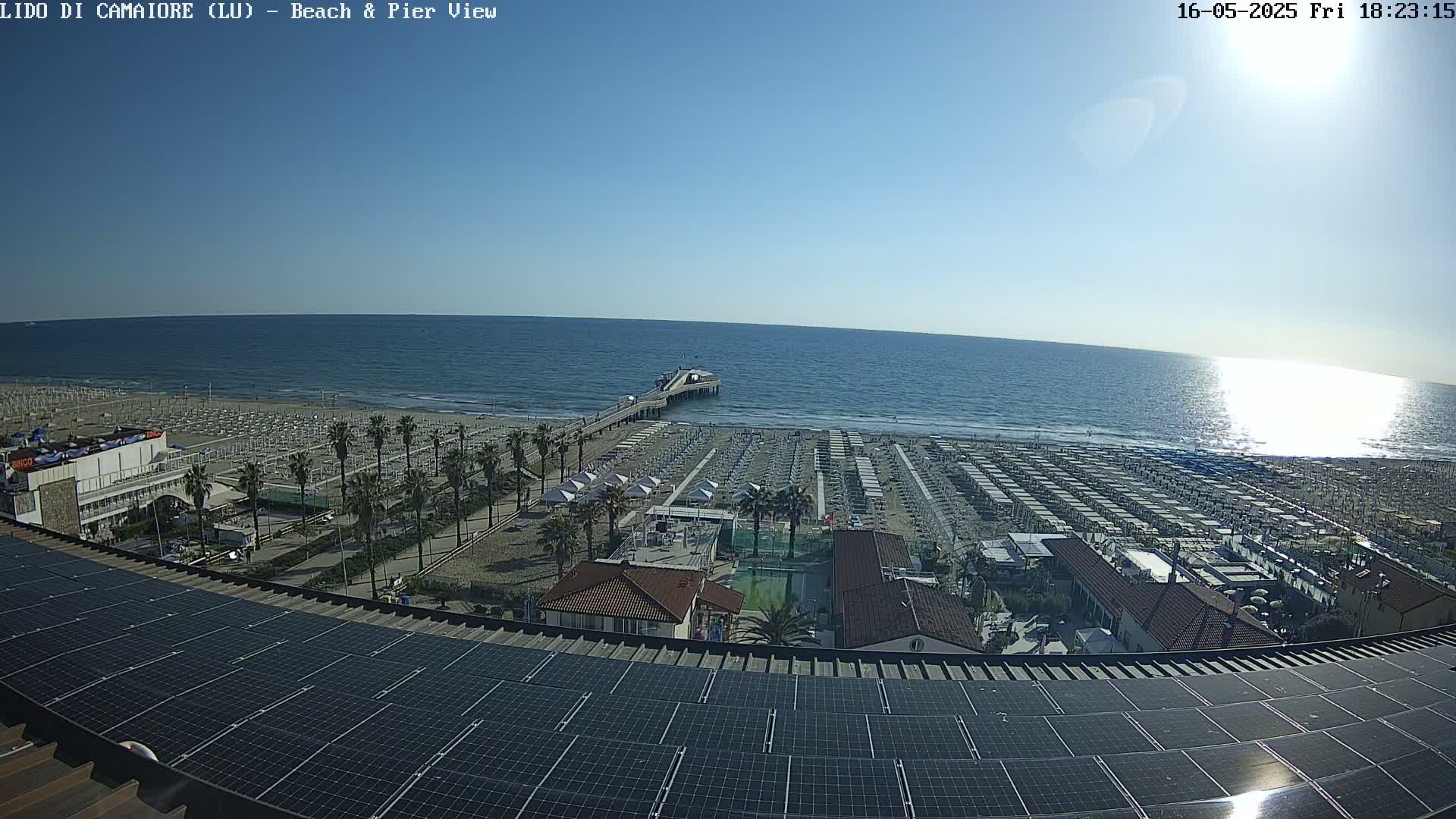 A beach with rows of beach umbrellas and a pier extending into a calm, sunny ocean is viewed from above a rooftop covered in solar panels.