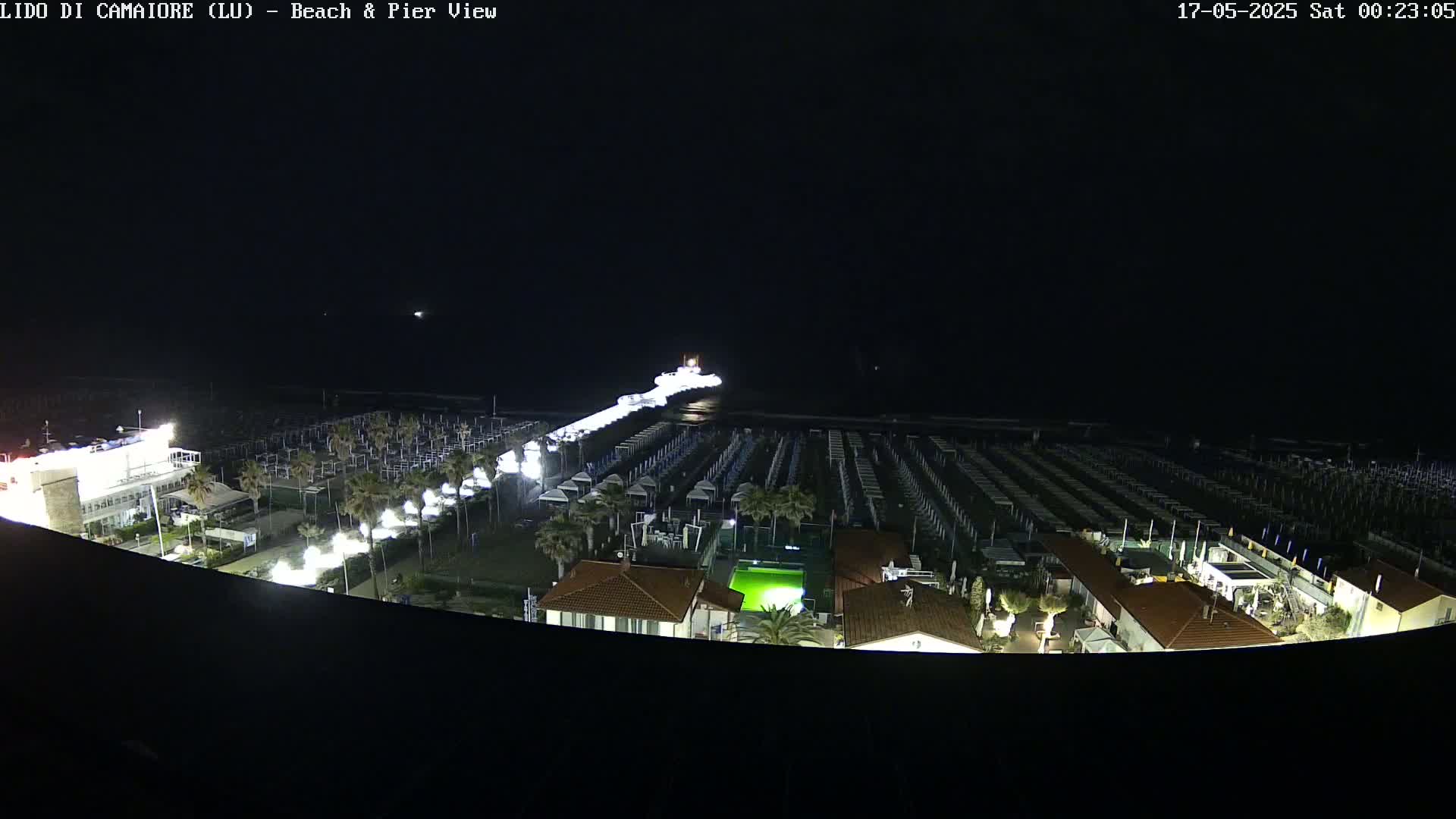 A nighttime aerial view shows a beach with rows of closed beach umbrellas, a pier extending into the dark ocean, and buildings with lights illuminating the scene.