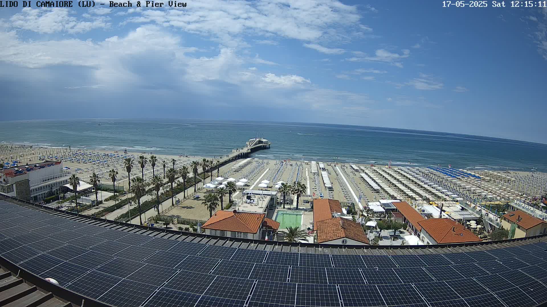 A sunny day at a beach with numerous umbrellas and a pier extending into a calm ocean, viewed from above a rooftop covered in solar panels.