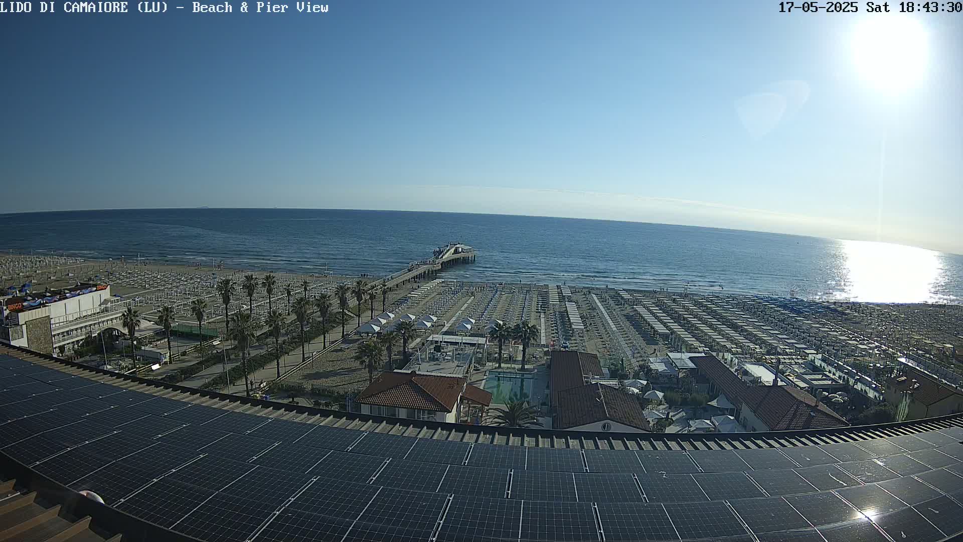A sunny, high-angle view shows a beach with rows of umbrellas and a pier extending into a calm ocean, all observed from a rooftop covered in solar panels.